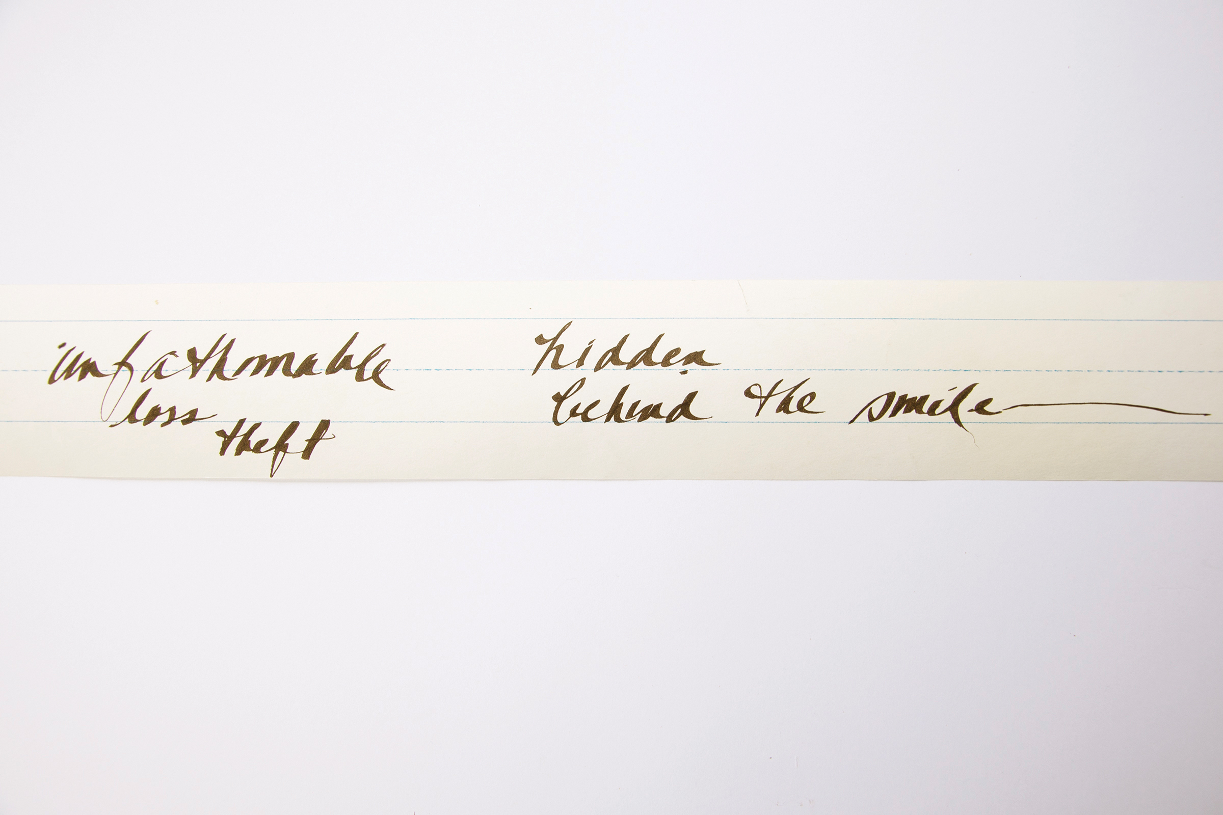 A narrow piece of white, lined paper with black, handwritten script that reads either unfathomable loss theft hidden behind the smile or unfathomable hidden loss behind the smile theft.