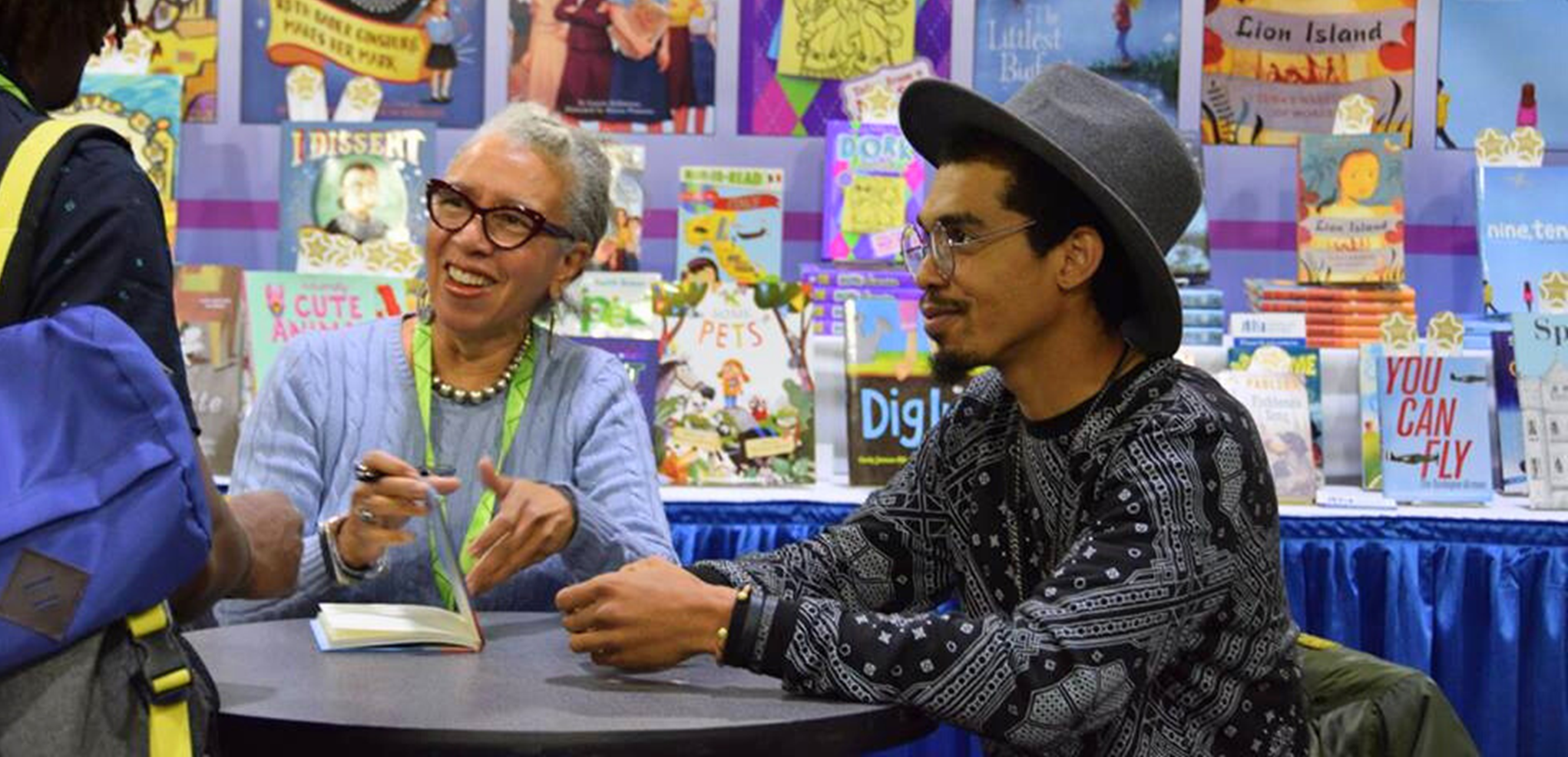 Carole Boston Weatherford and her son Jeffery Boston Weatherford seated at a table with colorful children's books on a wall behind them.