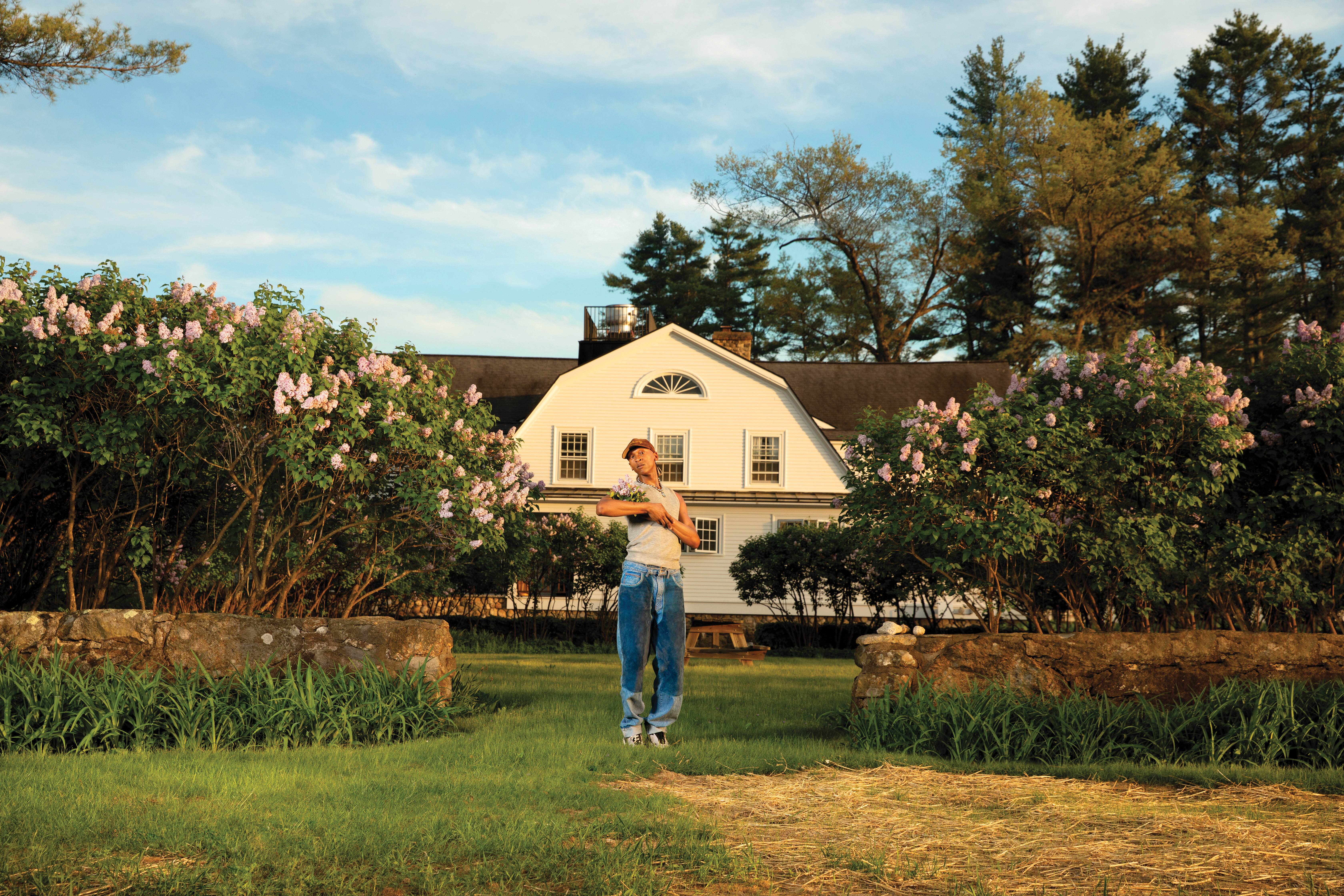 Golden wears jeans, a light gray sleeveless shirt, and a baseball cap and holds a bouquet of flowers. Lilac bushes are on either side of Golden and a large white house is in the background.