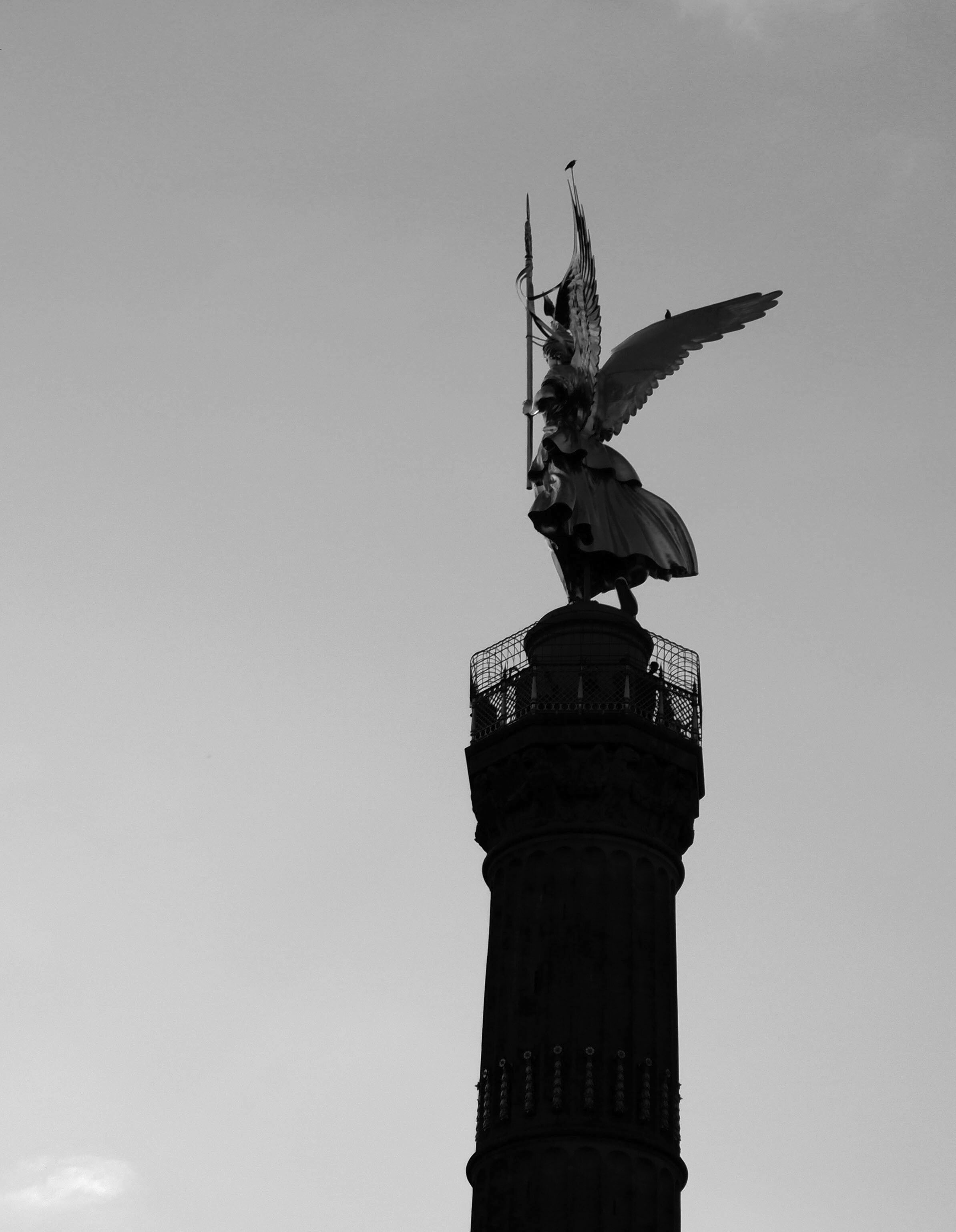 The statue of Victoria on the Victory Column in Berlin. The monument almost forms a silhouette against the sky. Victoria is winged and a couple of birds are sitting on the tips of the wings.