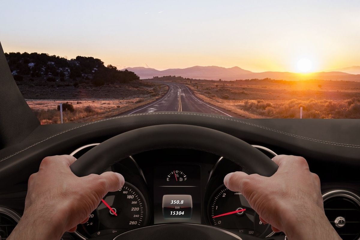 Hands gripping a car steering wheel driving down a dessert mountain road at sunset