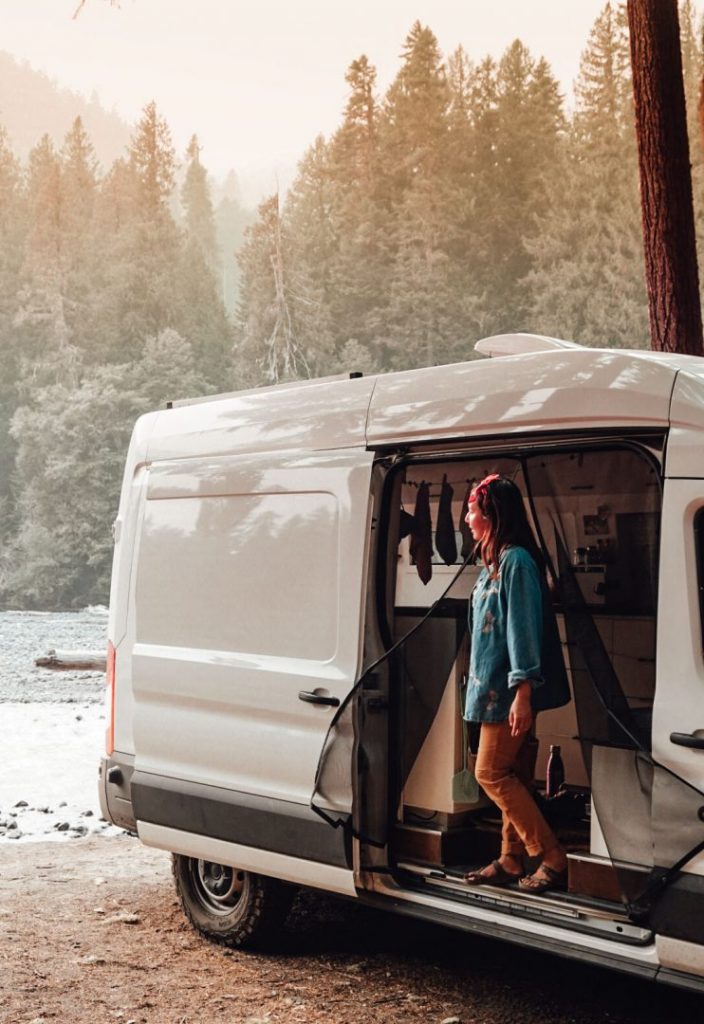 Woman stands in the open door of her camper van in a forest setting