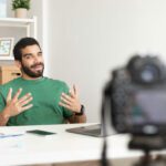 Glad Young Arabic Man Blogger At Table With Laptop Working Remotely, Shooting Vlog, Online Lesson