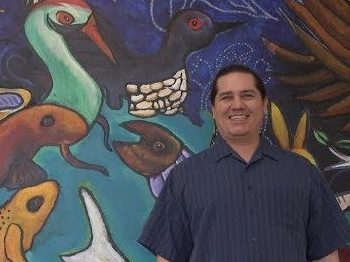 Teacher Reggie Cadotte (Lac Courte Oreilles Band of Lake Superior Chippewa) stands in front of a mural