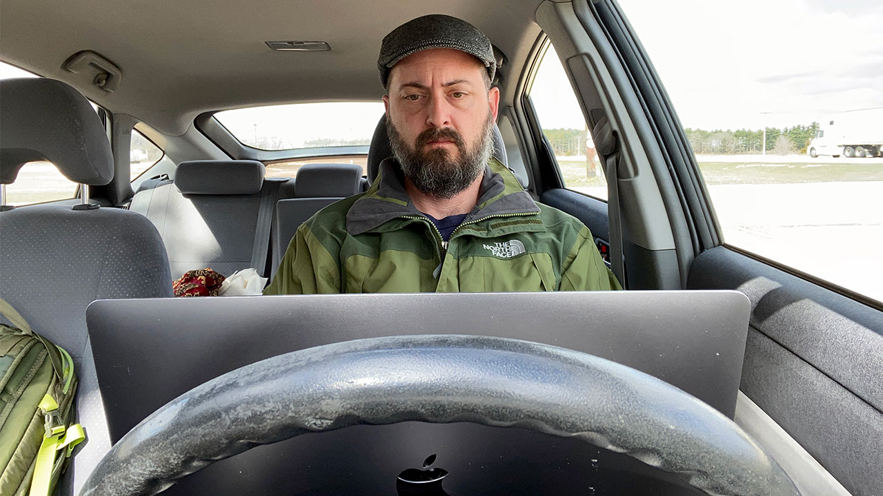 Man sitting behind steering wheel of car with an open laptop computer